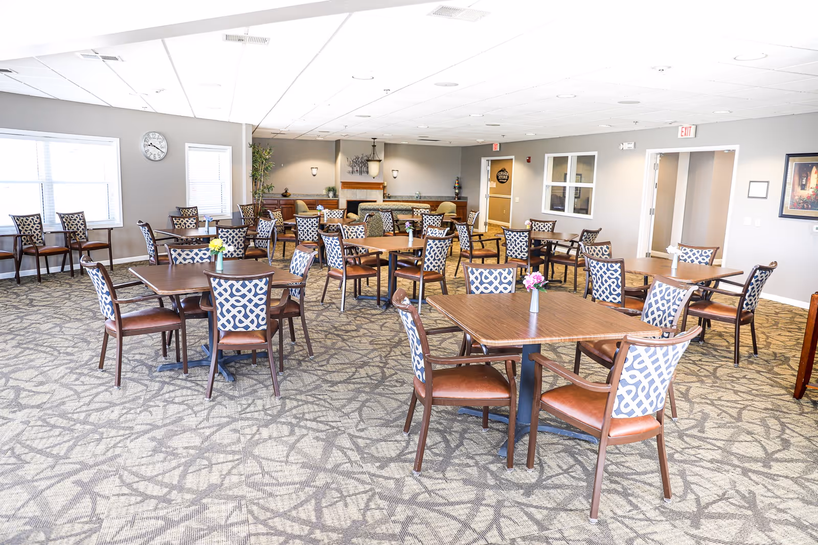 Spacious communal dining room with multiple square wooden tables and patterned chairs, small floral centerpieces, and large windows.