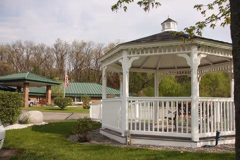 White wooden gazebo with a bench inside situated on a grassy area near a driveway. In the background, there is a building with a green metal roof, an American flag on a flagpole, and trees with sparse leaves.
