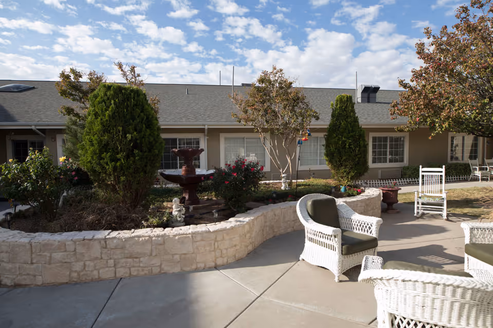 Outdoor patio area at a senior living facility with white wicker chairs and a stone planter containing small trees and bushes. A two-tiered water fountain is centered in the planter. The building with windows and a gray roof is visible in the background under a partly cloudy sky.
