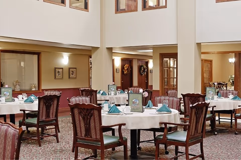 A dining room in a senior living facility with round tables covered in white tablecloths, each set with teal napkins, glasses, and menus. The room features wooden chairs with patterned upholstery, carpeted floors, and warm lighting. There are windows and doors with wooden frames, and decorative wreaths on some doors in the background.