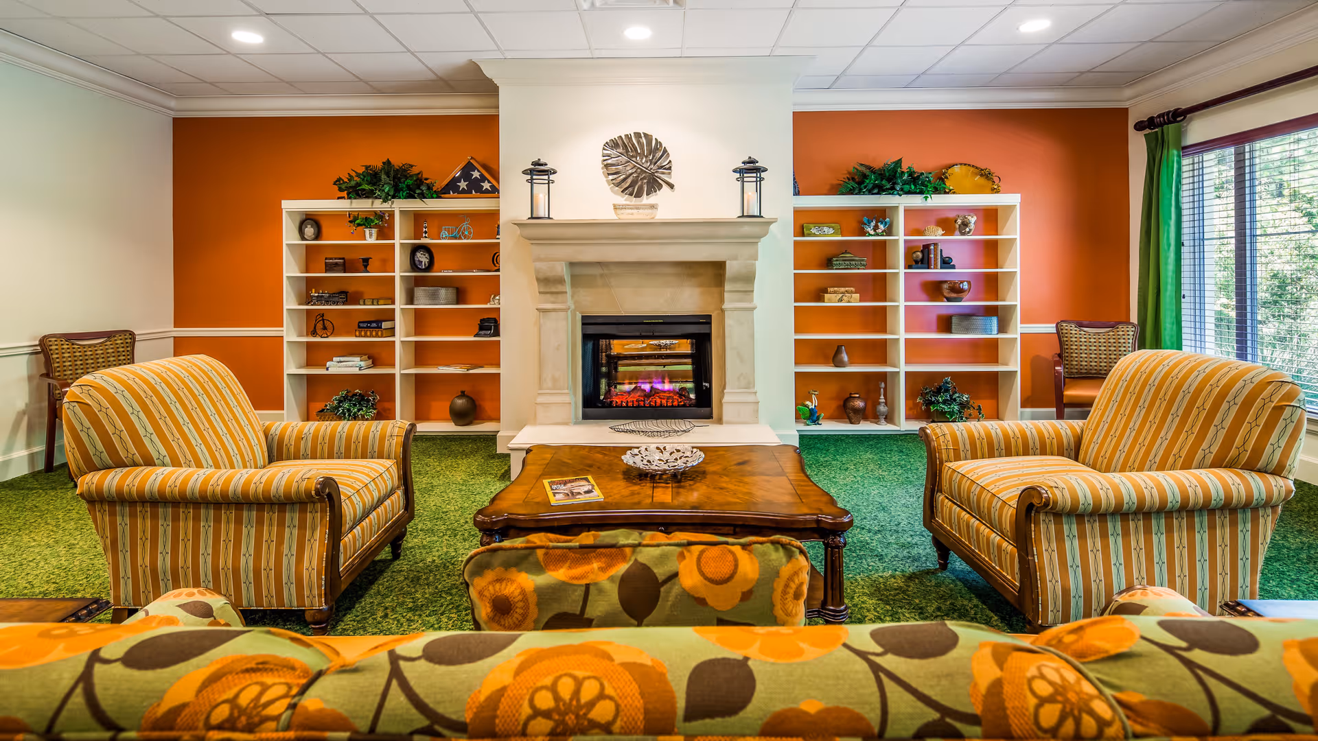 A cozy living room area with two striped armchairs facing a wooden coffee table and a fireplace. The room has green carpet, orange accent walls with white shelves holding decorative items, and a large window with green curtains letting in natural light.