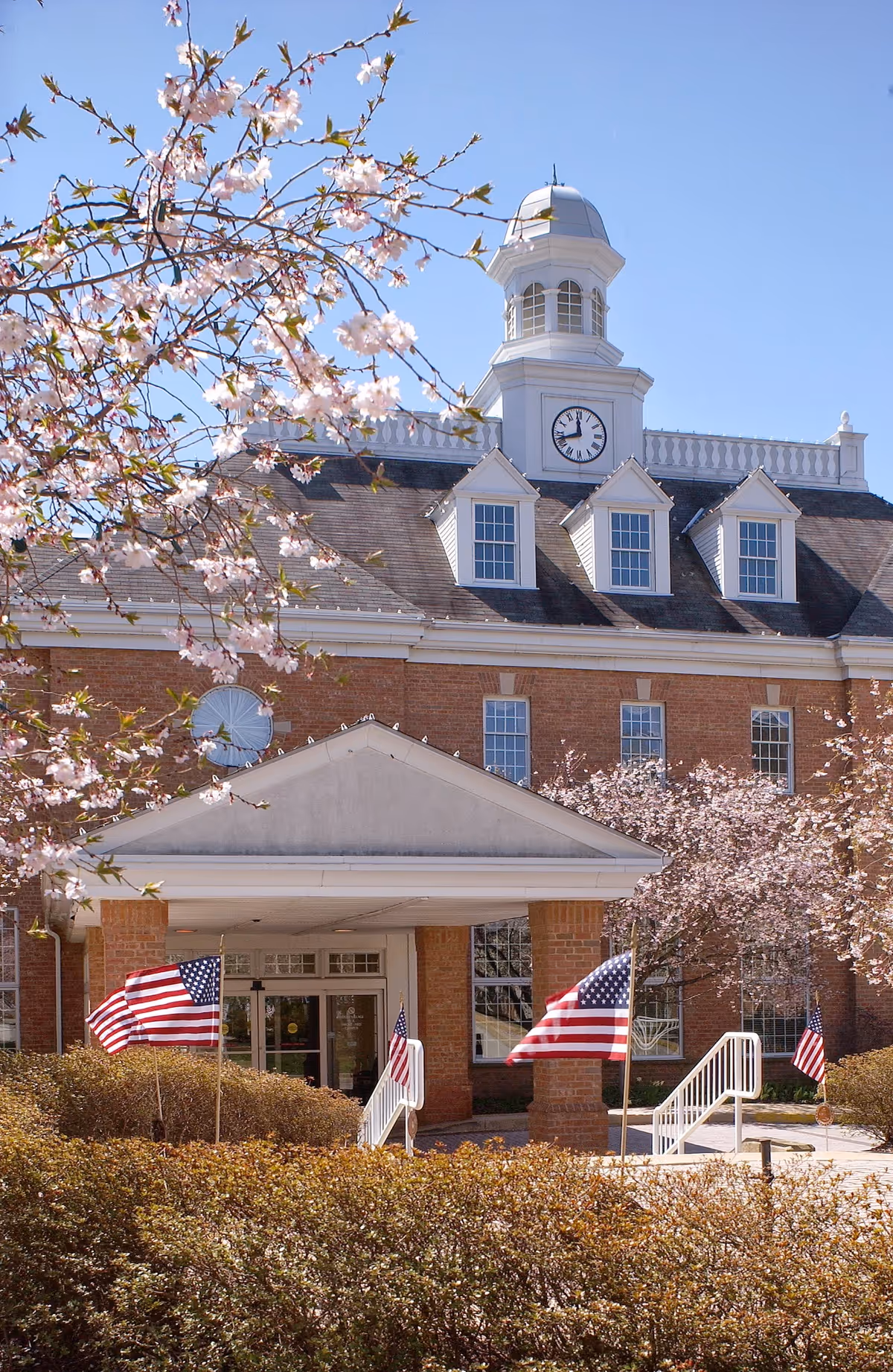 Front entrance of a brick retirement community building with a clock tower, American flags, and flowering trees.