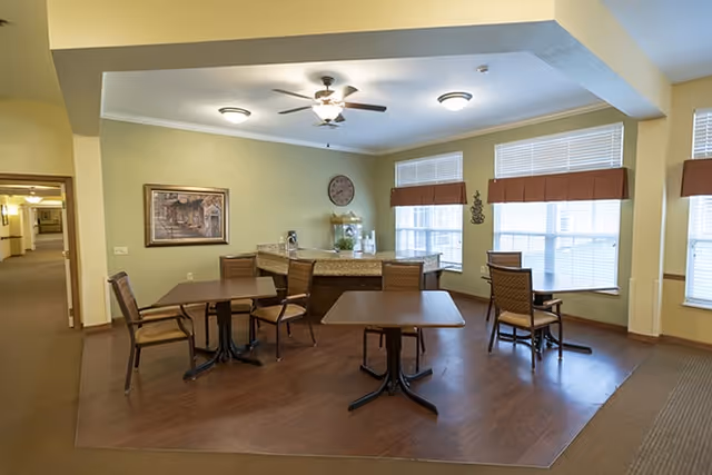 A common area in a senior living facility with several tables and chairs arranged on a wooden floor. The room has large windows with blinds, a ceiling fan with lights, a clock on the wall, and a counter with a coffee machine and water dispenser. The walls are painted light green and beige, and there is a framed picture hanging on the wall.