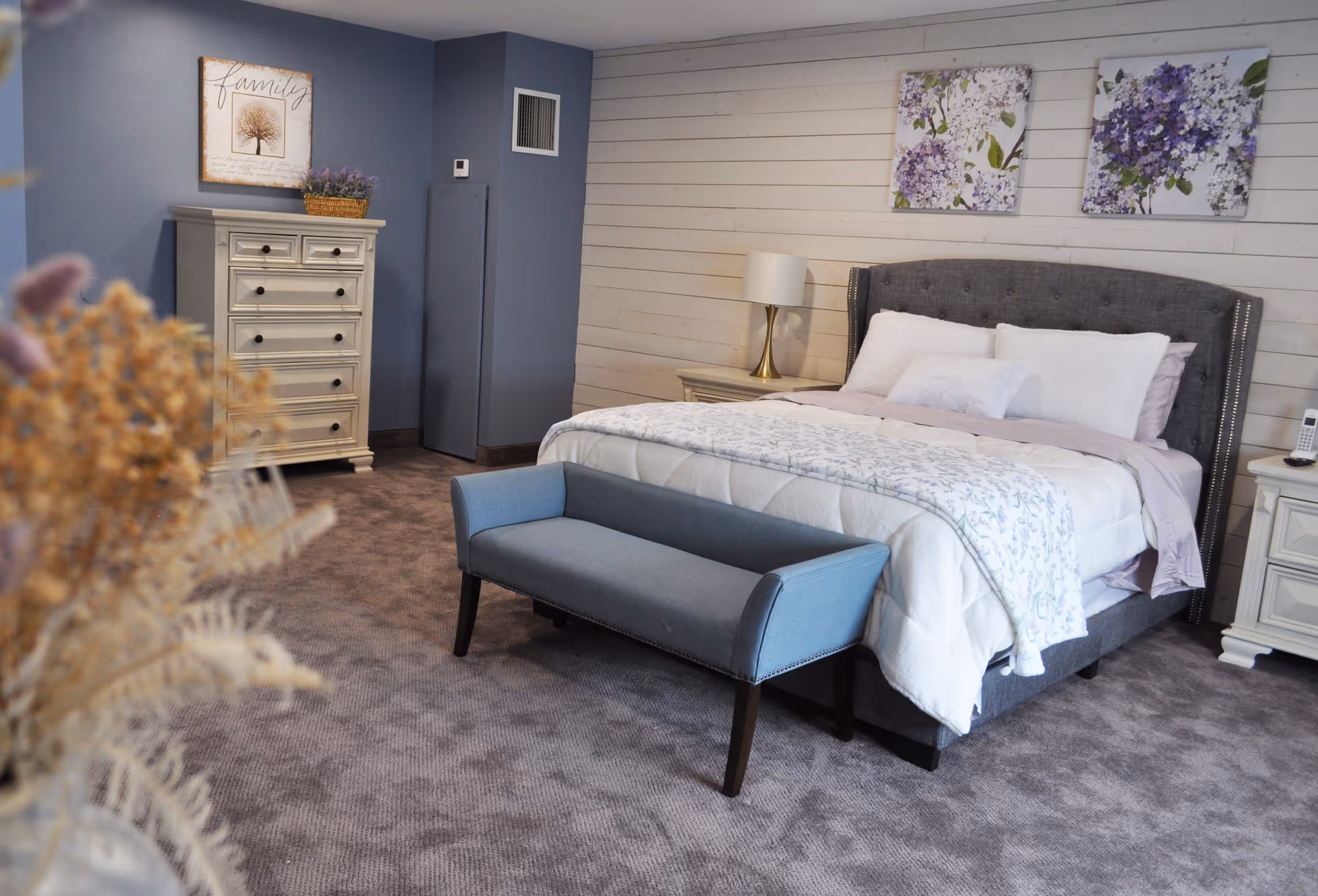 A cozy bedroom with a large bed featuring white and light purple bedding, a gray upholstered headboard, and a blue bench at the foot of the bed. There are two nightstands with lamps on either side of the bed, a white dresser with drawers against a blue wall, and two floral paintings hanging above the bed. The room has carpeted flooring and a decorative framed picture on the wall.