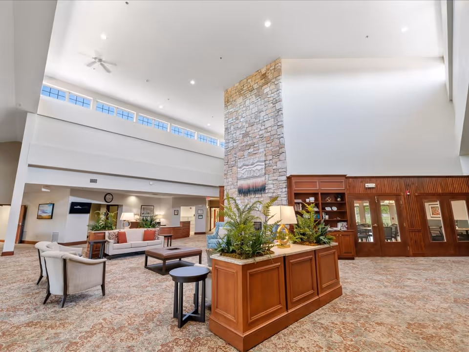 Spacious senior living facility common area with high ceilings, a large stone fireplace, comfortable seating including sofas and armchairs, wooden cabinetry with plants and lamps, and large windows near the ceiling allowing natural light.