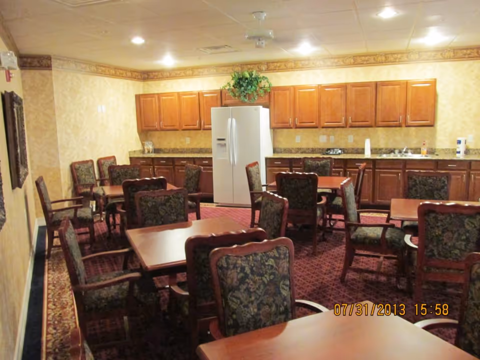 Interior view of a dining room with multiple wooden tables and upholstered chairs arranged on a patterned carpet. The back wall features wooden cabinets, a white refrigerator, and a countertop with a sink and various items. The ceiling has recessed lighting and a ceiling fan with a plant hanging above the refrigerator.