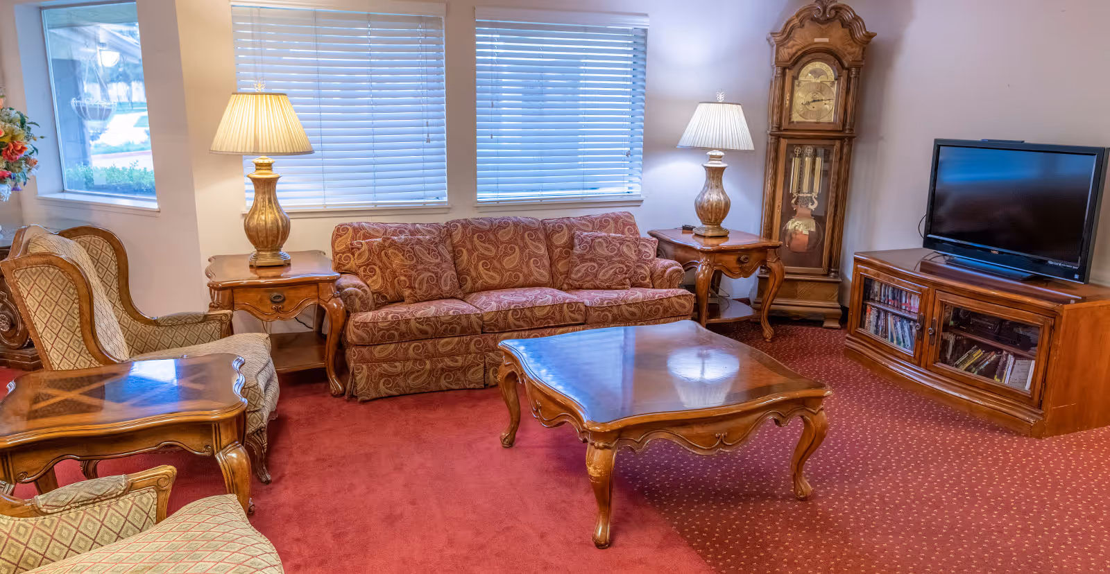 A cozy living room with a patterned red sofa, two matching armchairs, two wooden side tables with lamps, a wooden coffee table, a grandfather clock, and a TV on a wooden cabinet. The room has red carpet and two windows with blinds.