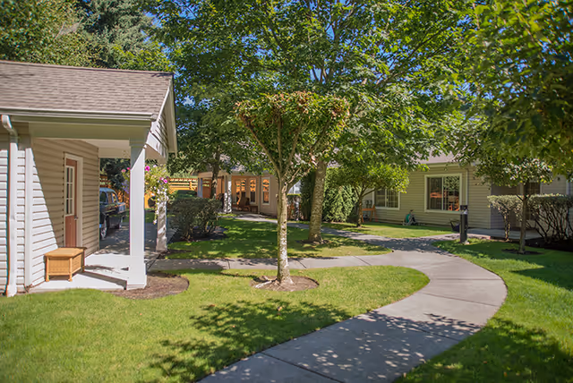 A sunny outdoor courtyard area with a curved concrete pathway surrounded by green grass, trees, and shrubs. Several single-story buildings with light-colored siding and windows are visible around the courtyard.