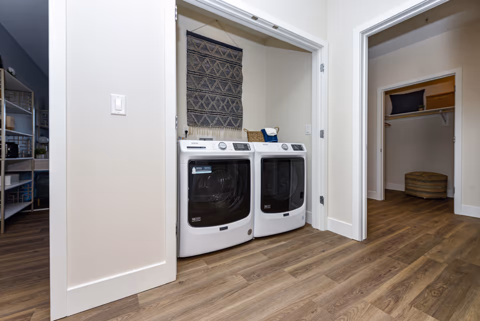A laundry area with a white front-loading washing machine and dryer side by side in a small closet space. Above the machines, there is a decorative woven wall hanging. The floor is wood, and adjacent rooms with open doors are visible, including a closet with shelves and storage items.