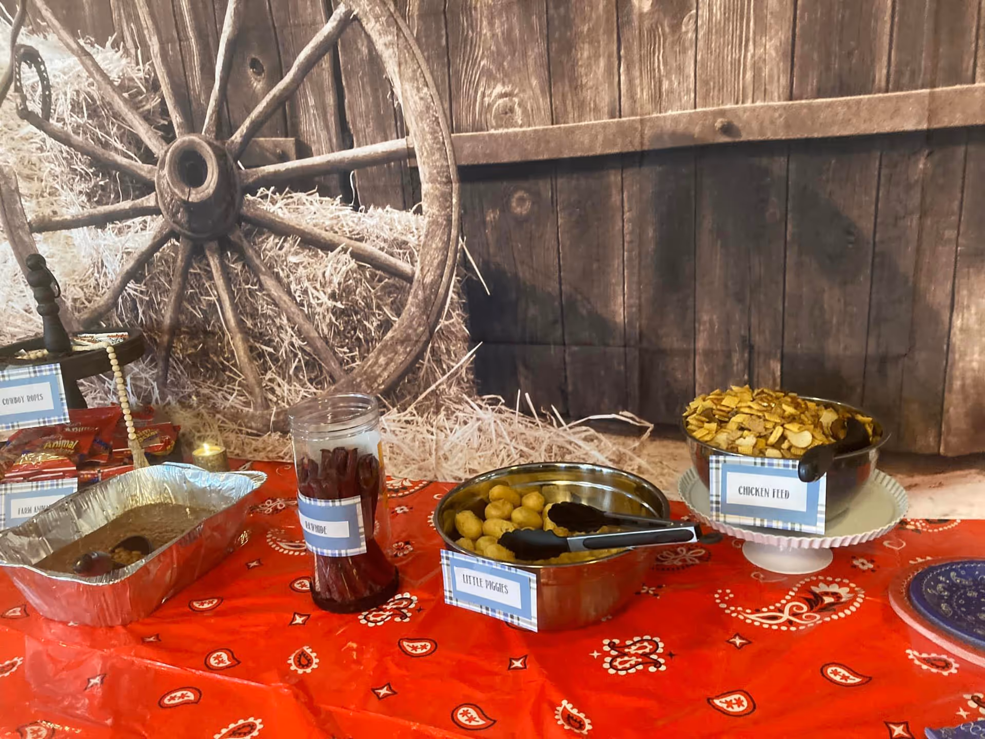 Rustic dining table with red tablecloth, hay bales, and wagon wheel backdrop, labeled bowls of snacks.