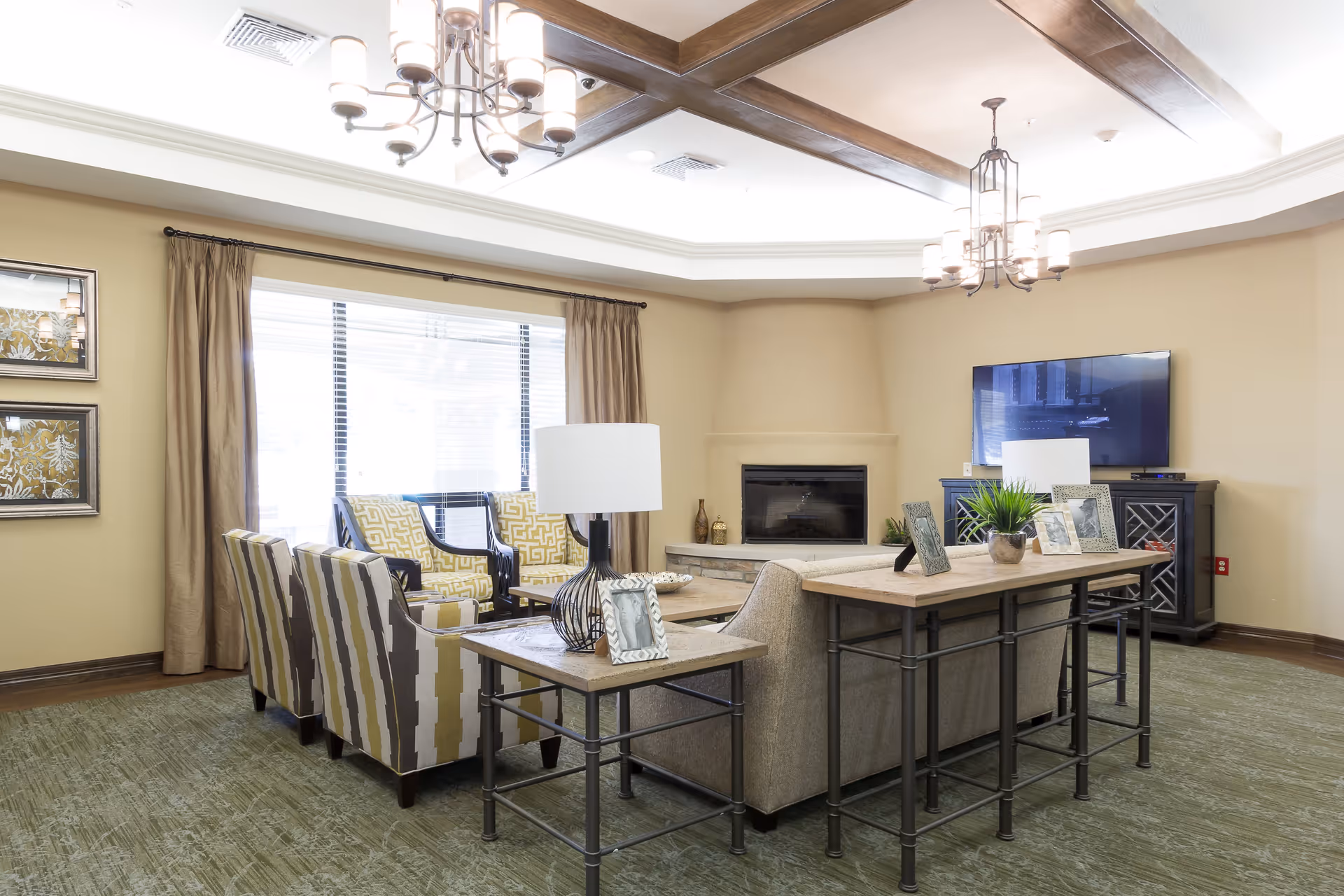 A bright and spacious senior living facility common area with beige walls and green carpet. The room features a seating arrangement with patterned armchairs and a sofa around a wooden coffee table. Behind the sofa is a console table with framed photos and a potted plant. A flat-screen TV is mounted on a dark wooden cabinet, and a fireplace is built into the far wall. The ceiling has wooden beams and two modern chandeliers. Large windows with beige curtains allow natural light to fill the room.
