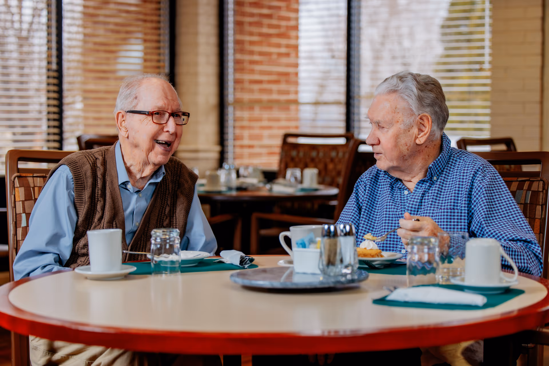 Two elderly men sitting at a round table in a dining area, engaged in conversation. One man is wearing glasses, a blue shirt, and a brown vest, while the other is wearing a blue checkered shirt and eating dessert. The background shows windows with blinds and other tables and chairs.
