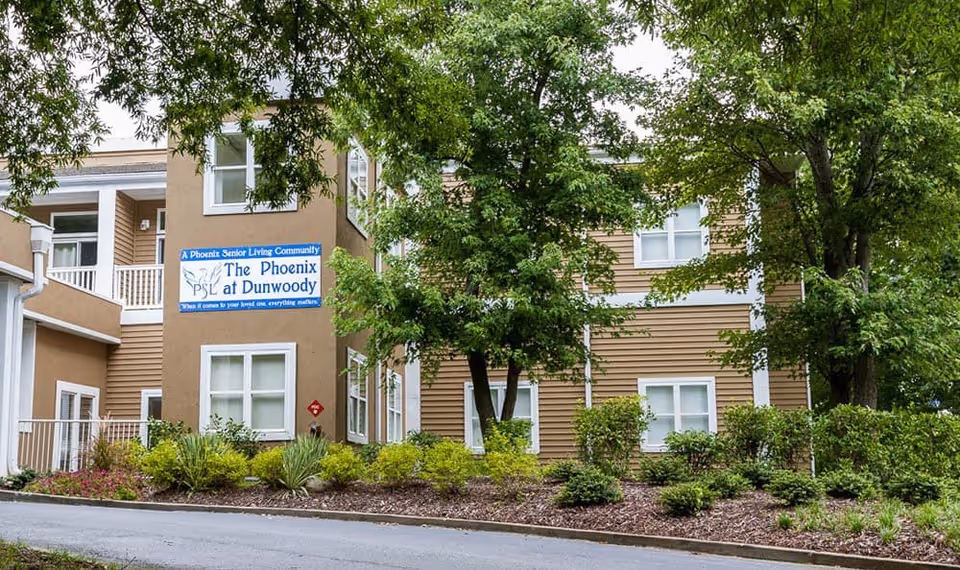 Exterior view of The Phoenix at Dunwoody senior living community building surrounded by trees and landscaped bushes with a sign displaying the facility name.