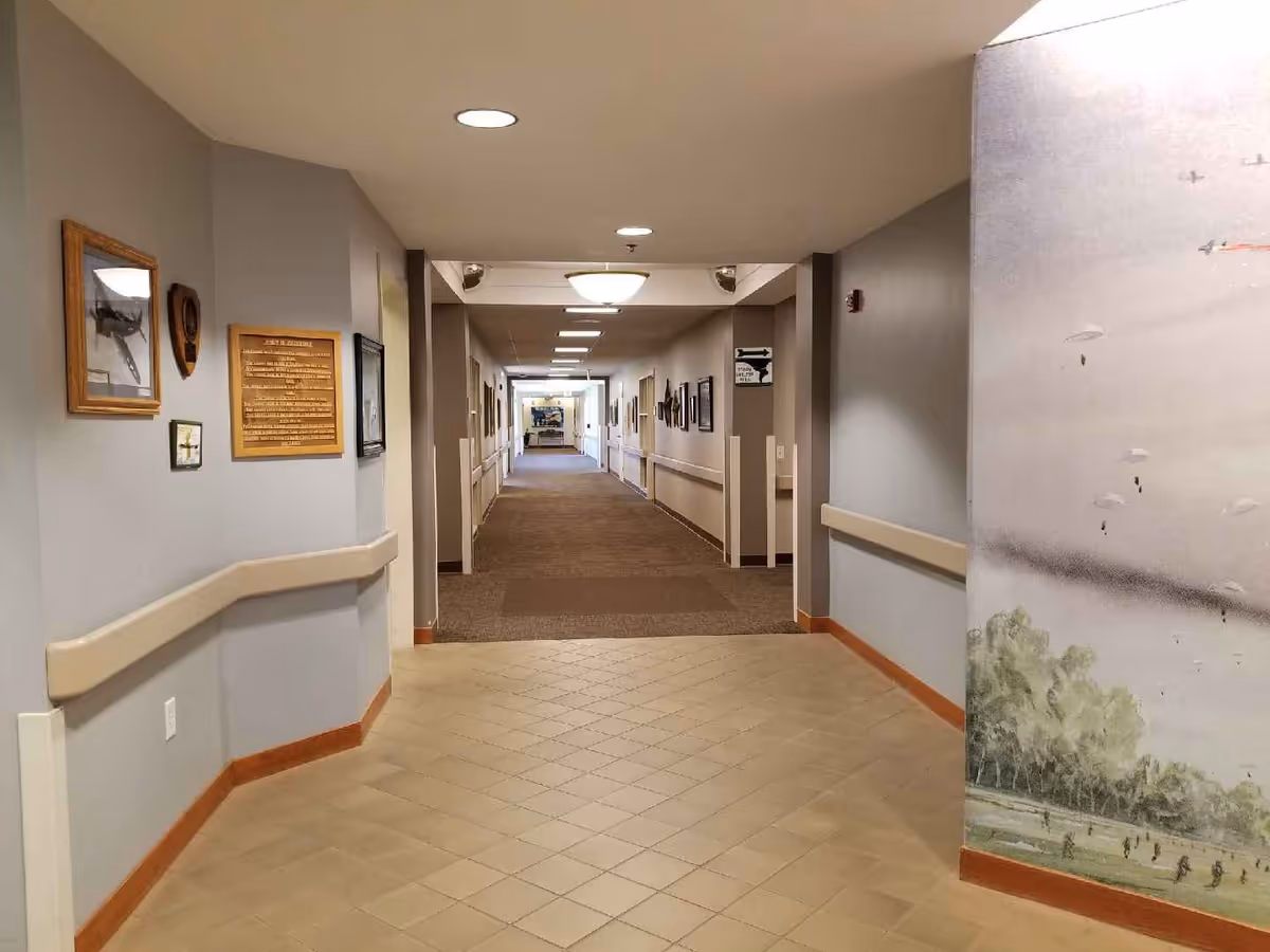 Long well-lit interior hallway with a tiled entry, carpeted corridor, wall handrails, and framed pictures and plaques.