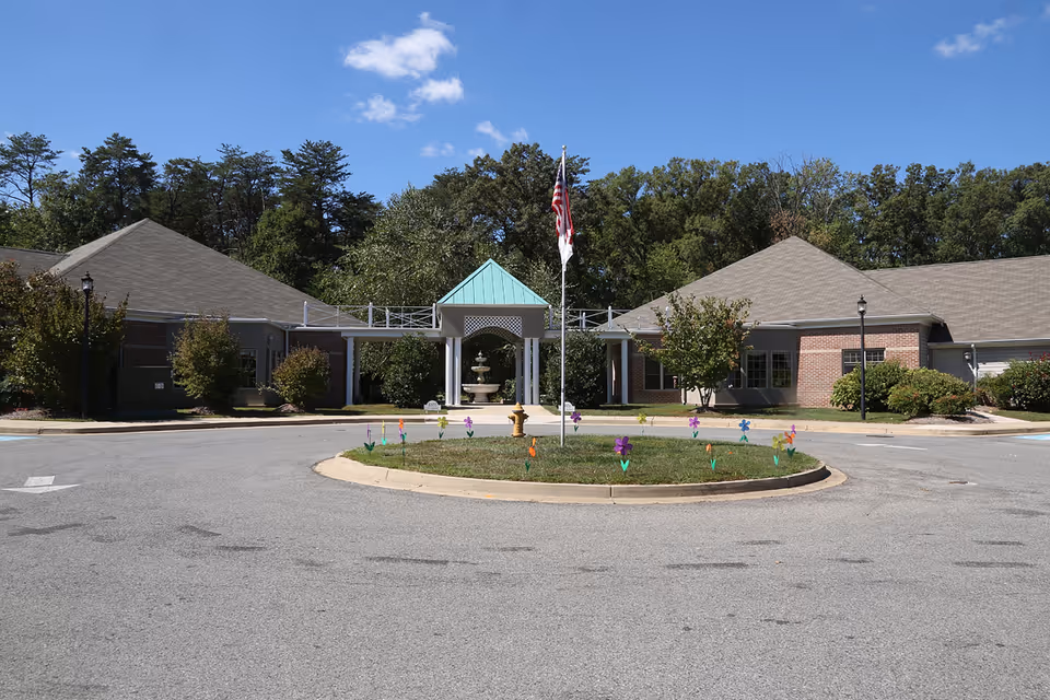 Front exterior view of Fenwick Landing Senior Care Community building with a circular driveway, a flagpole with an American flag, a small grassy island decorated with colorful flower-shaped decorations, and trees in the background under a clear blue sky.