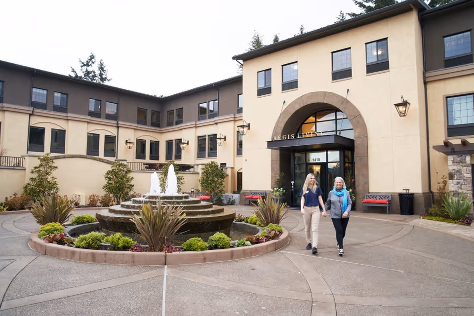 Two women walking and holding hands outside the entrance of Aegis Living Ravenna, a multi-story senior living facility with a central fountain and landscaped plants in the courtyard.