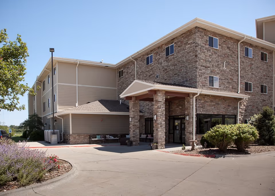 Three-story brick and siding senior living building with a covered main entrance, driveway, and landscaped shrubs.