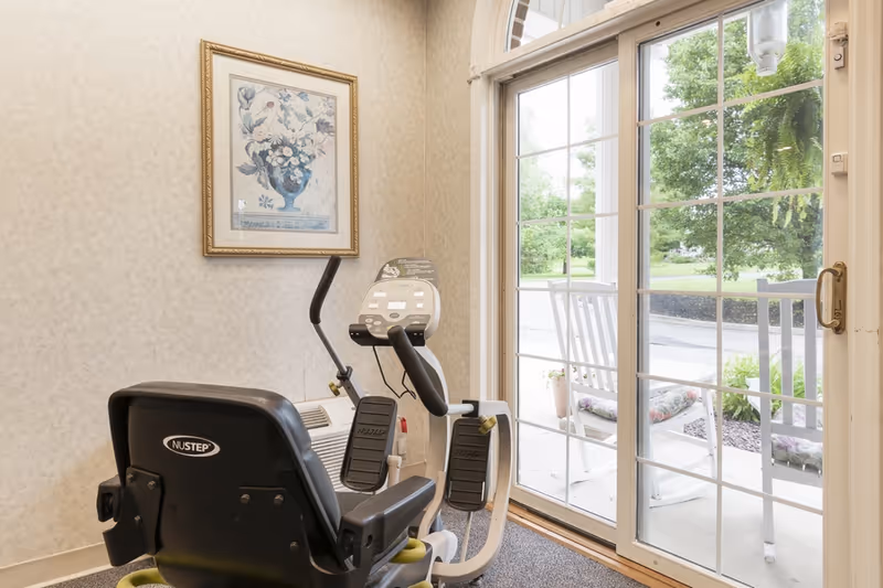 Recumbent exercise bike in a small interior room with a framed floral picture and sliding glass doors opening to a porch with rocking chairs.