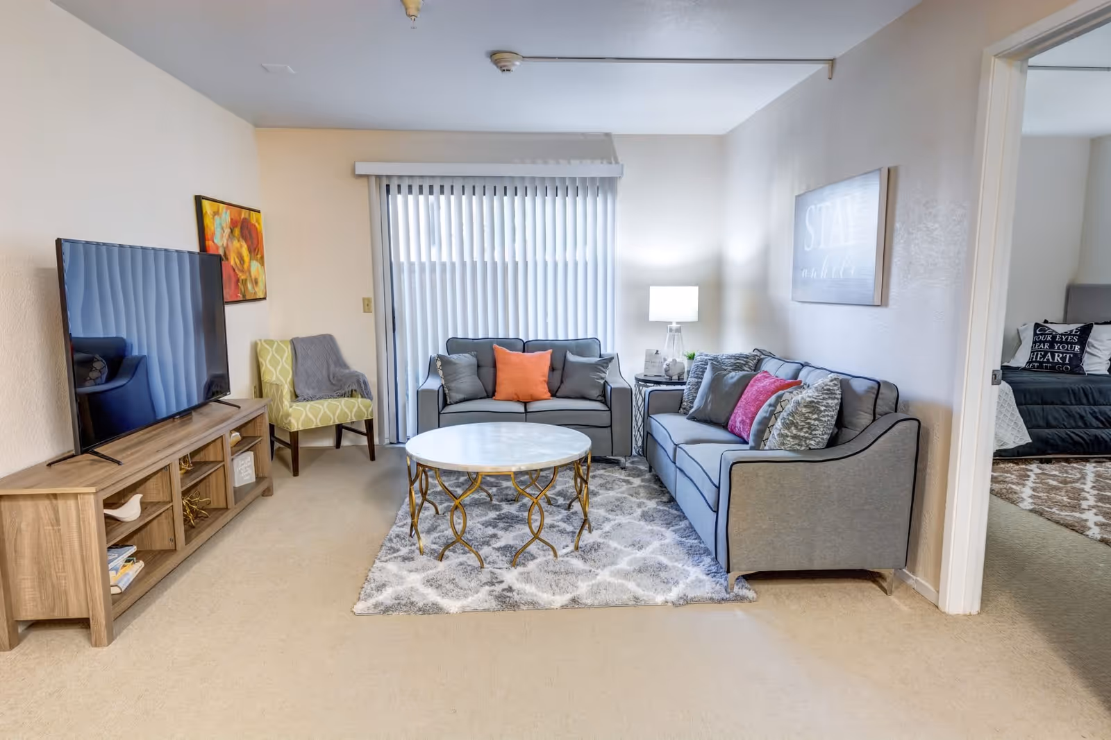 A cozy living room with two gray sofas adorned with colorful pillows, a round coffee table with a decorative gold base, a patterned area rug, a wooden TV stand with a large flat-screen TV, a green patterned armchair, a side table with a lamp, and vertical blinds covering a sliding glass door. An adjacent bedroom is partially visible through an open doorway.