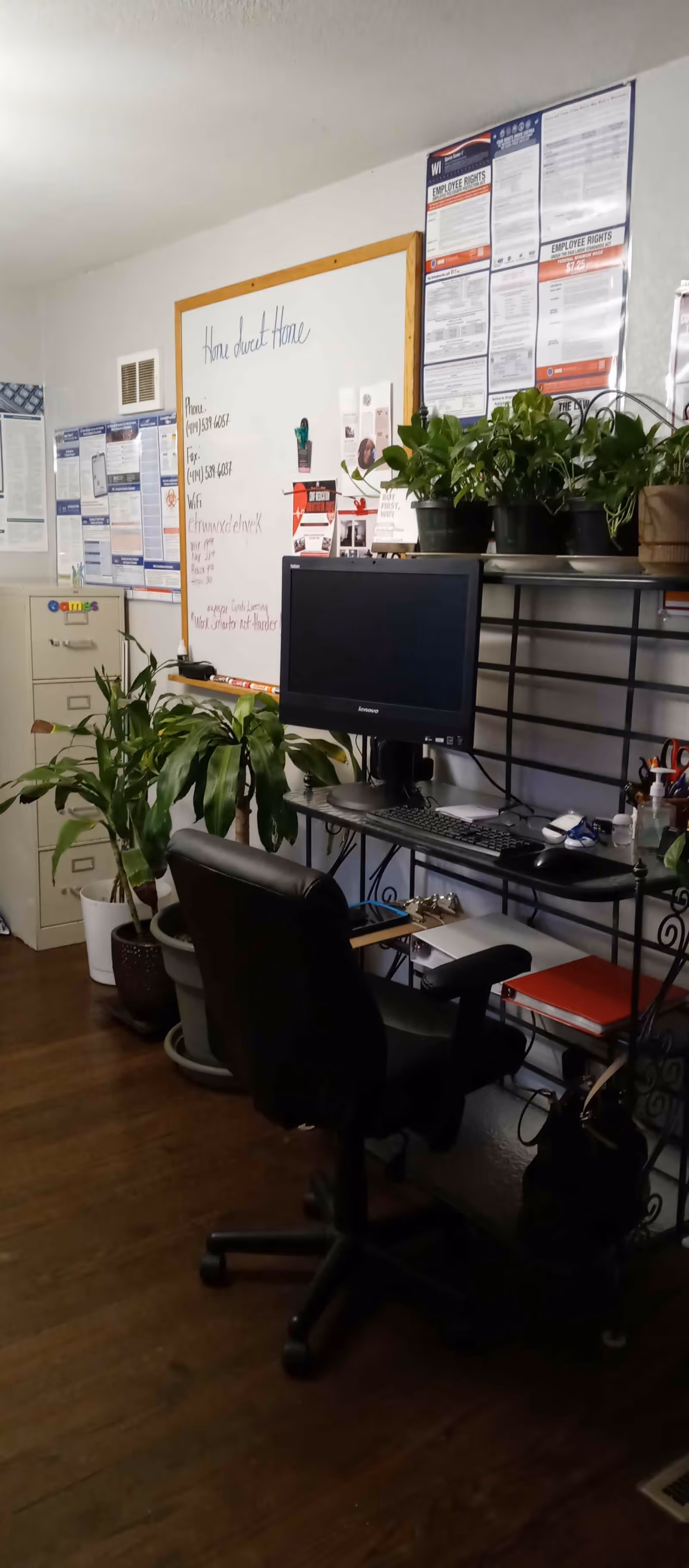 An office space with a black office chair in front of a desk holding a computer monitor, keyboard, and mouse. Several potted plants are placed on the desk and floor. A whiteboard on the wall displays handwritten notes and contact information for Home Sweet Home Assisted Living LLC. Various posters and notices are pinned on the wall above and beside the whiteboard. A filing cabinet is visible in the background.