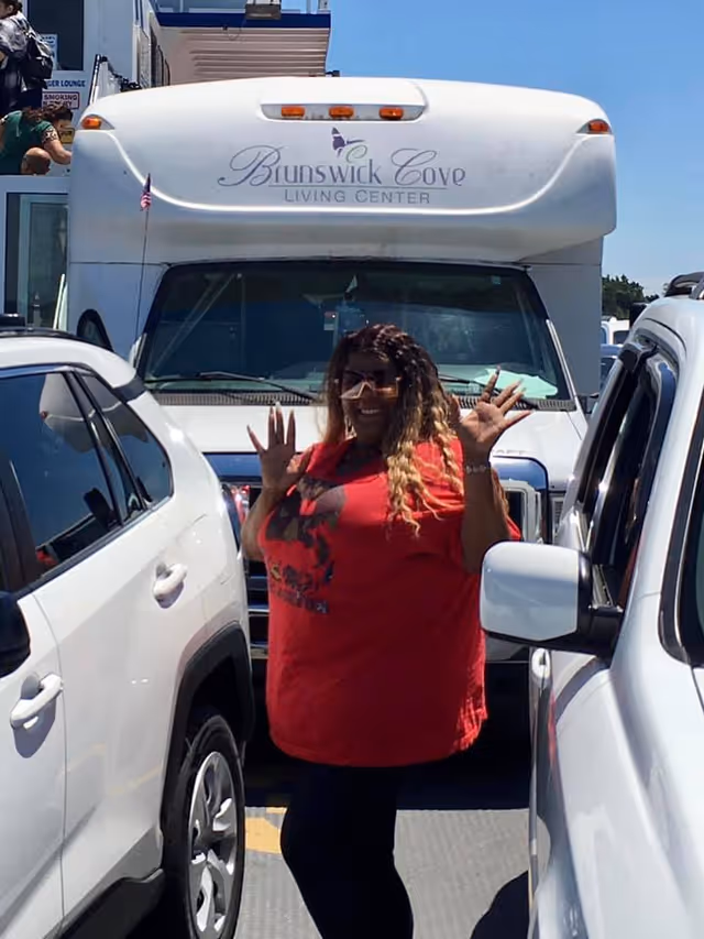 A woman wearing a red shirt and sunglasses stands between two white vehicles in a parking area, smiling and waving her hands. Behind her is a large white vehicle with the text 'Brunswick Cove Living Center' displayed on the front.