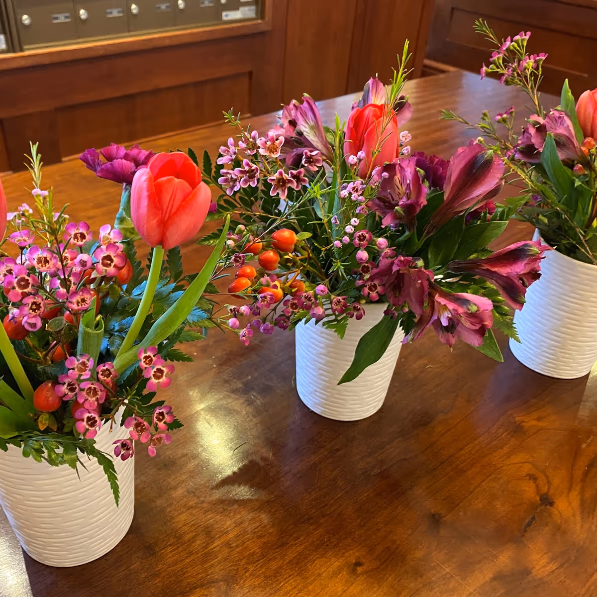 Three white textured vases with colorful floral arrangements including red tulips, purple flowers, and small pink blossoms placed on a polished wooden table with a wooden cabinet in the background.