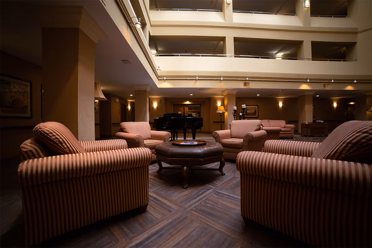 Dimly lit atrium lounge with striped armchairs arranged around a central ottoman and a piano in the background.
