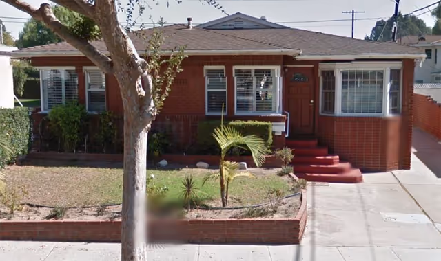 Front exterior view of a single-story brick house with a red door, white-framed windows, and a small front yard with some plants and a tree.