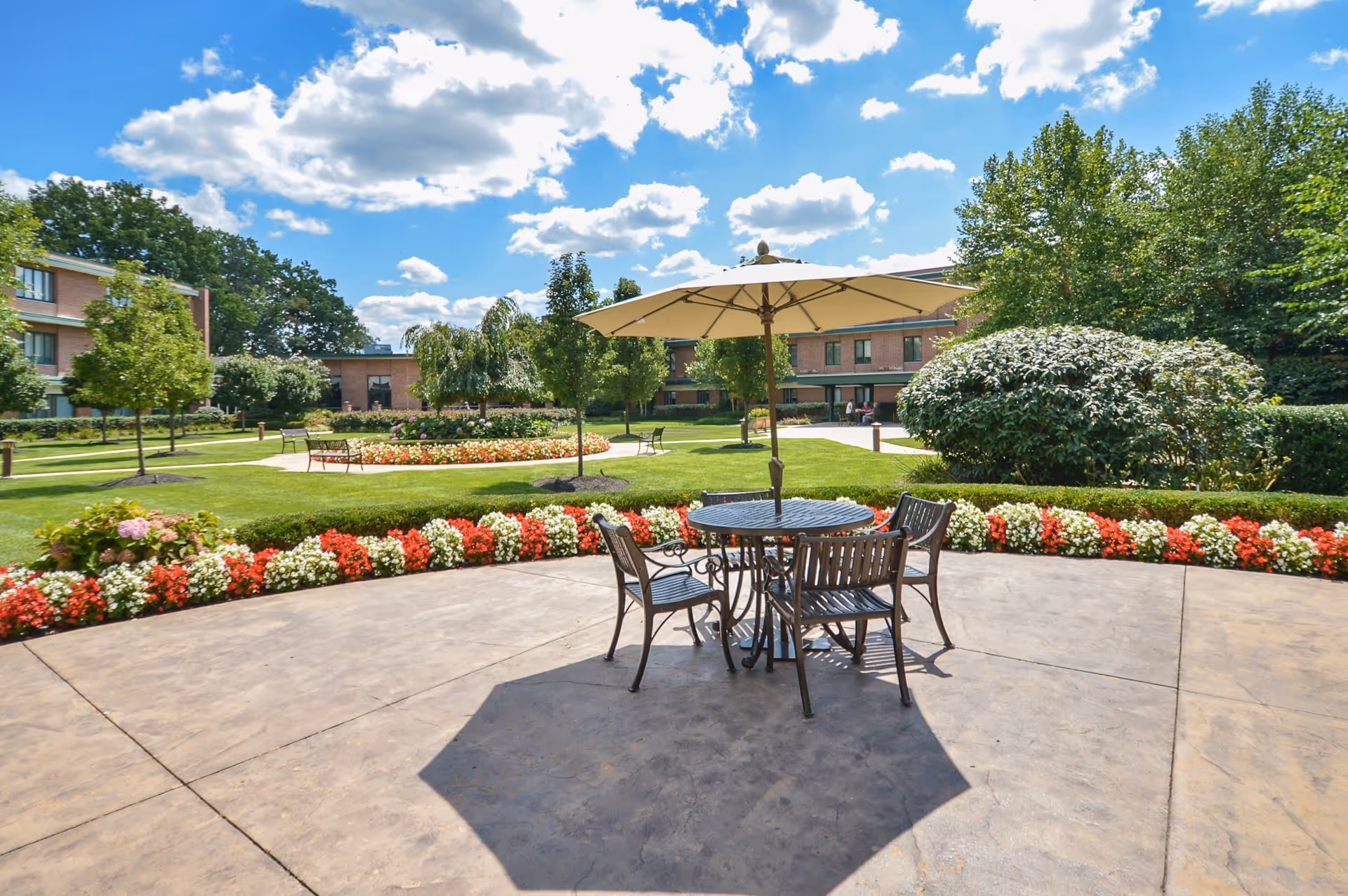 Outdoor patio area with a round metal table and four chairs under a large beige umbrella, surrounded by a well-maintained garden with colorful flowers, green grass, and trees. In the background, there is a two-story brick building under a partly cloudy blue sky.