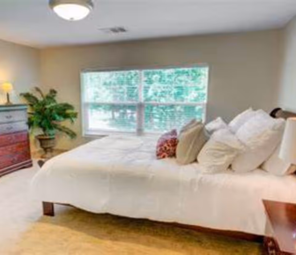 A bright bedroom with a large bed covered in white bedding and multiple pillows. There is a wooden dresser with a lamp and a potted plant next to it. A large window with blinds lets in natural light, and the walls are painted a light beige color.