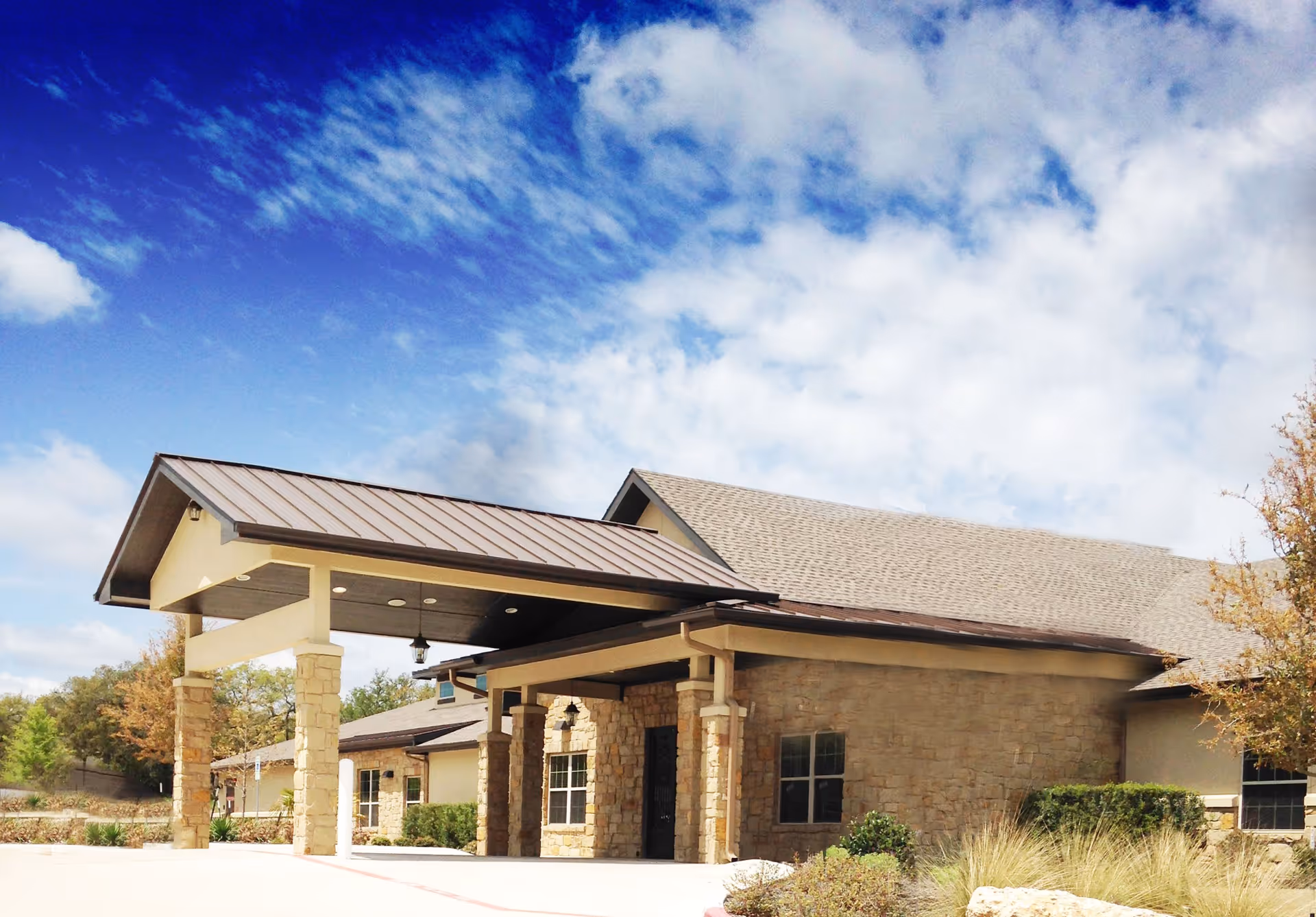 Exterior view of a single-story building with stone and beige walls, a covered entrance supported by stone pillars, and a sloped roof under a partly cloudy blue sky.