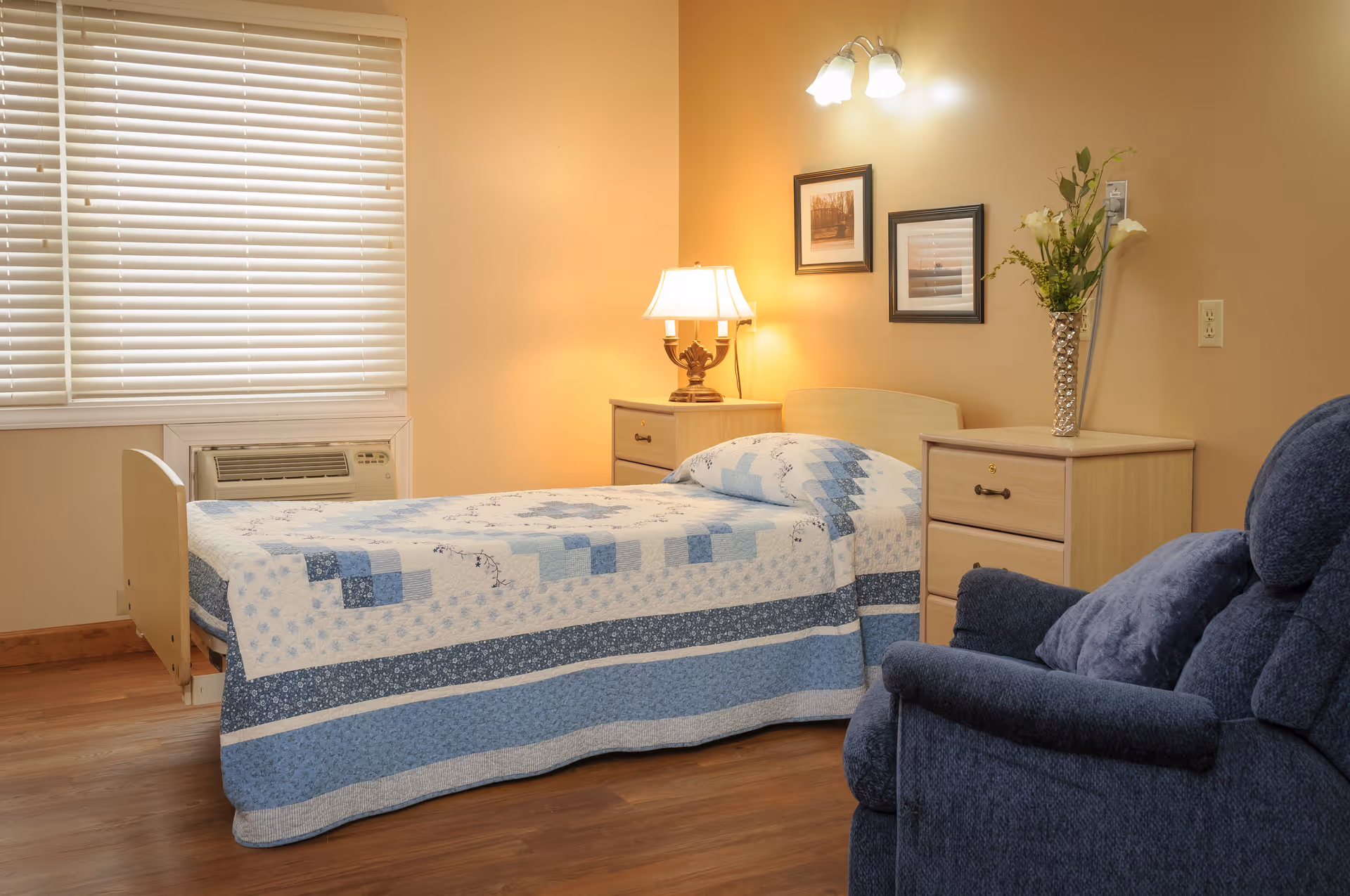 A cozy bedroom in a senior living facility featuring a single bed with a blue and white patchwork quilt, a wooden nightstand with a lamp, a wooden dresser with a vase of flowers, two framed pictures on the wall, a window with closed blinds, and a comfortable blue armchair.