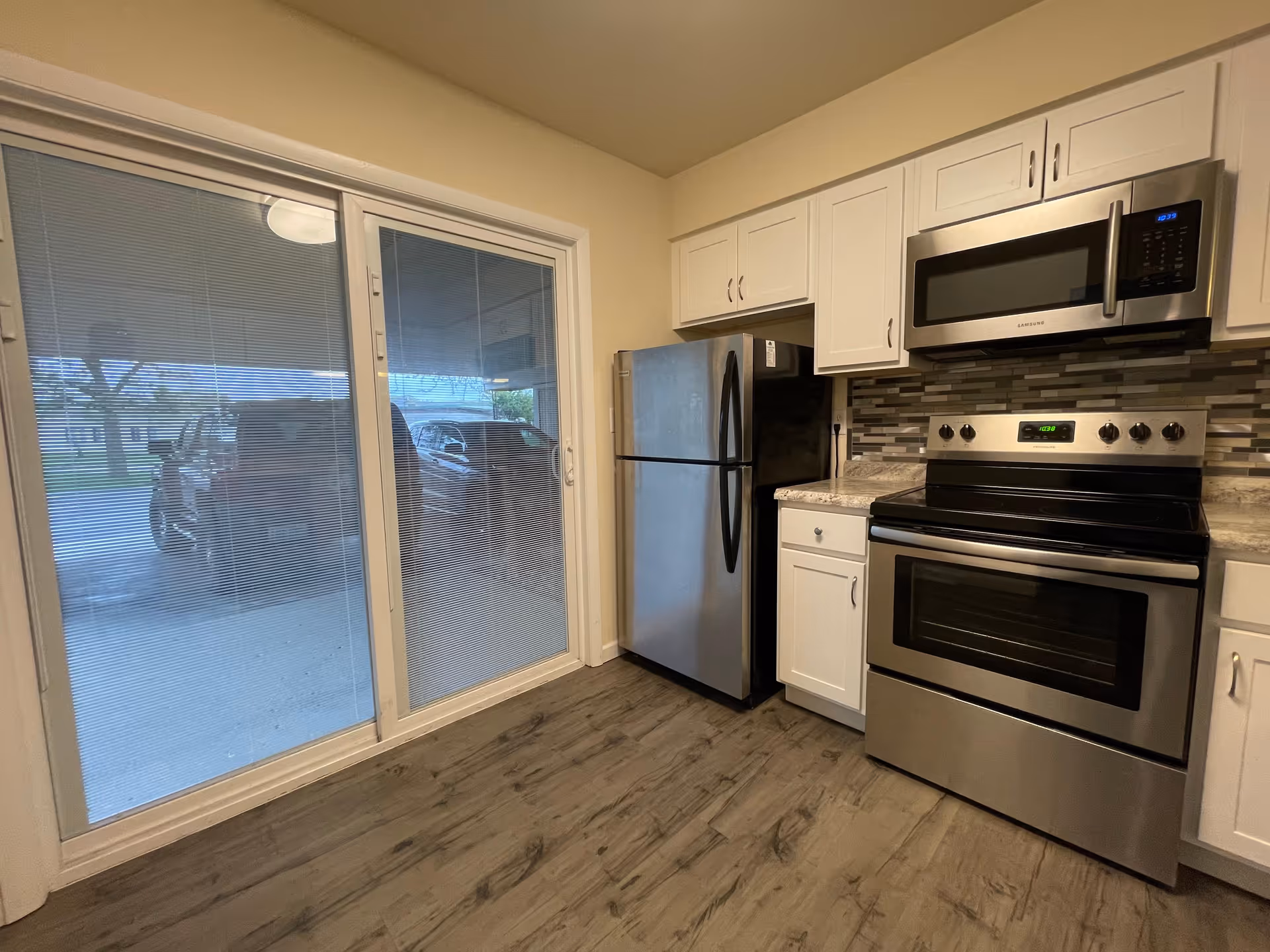 Modern kitchen with stainless steel refrigerator, oven and microwave, white cabinets and a sliding glass door showing parked cars outside.
