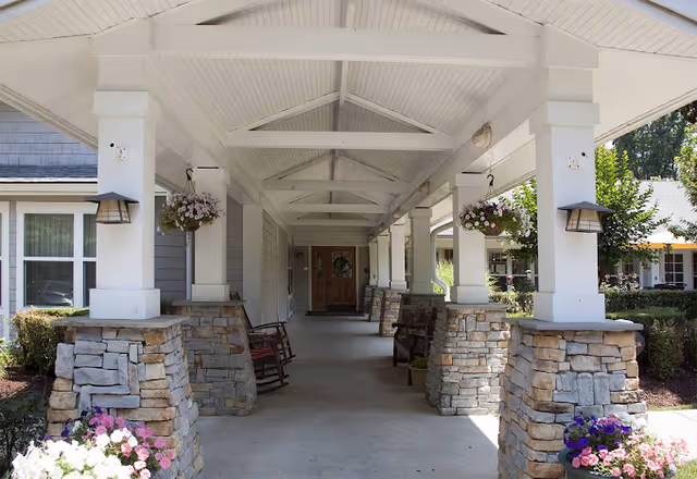 Covered entrance walkway with stone columns, hanging flower baskets, rocking chairs, and wooden double doors at the end.
