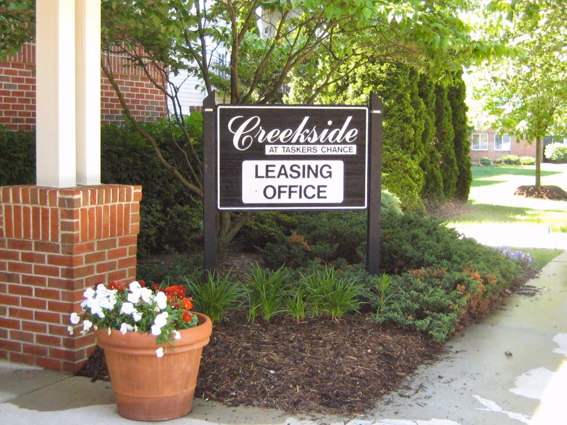 A wooden sign reading 'Creekside at Taskers Chance LEASING OFFICE' at a landscaped entrance with a brick column and potted flowers.