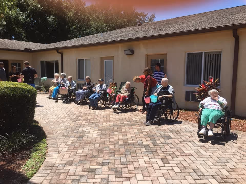 A group of elderly people, many in wheelchairs, sitting and standing outside on a paved patio area next to a single-story building. Some caregivers or staff members are standing and interacting with the residents. The scene is sunny with greenery and plants around the patio.