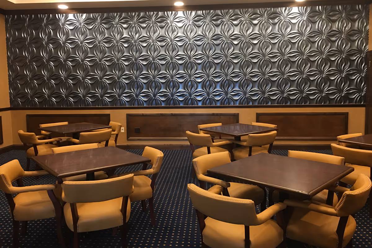 Interior view of a dining area with several square wooden tables surrounded by cushioned beige chairs. The room features a dark patterned carpet and a decorative wall panel with a textured, metallic floral design.