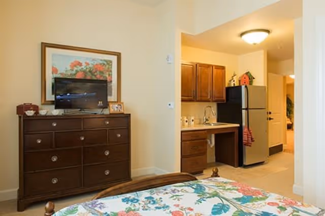Interior view of a senior living facility room showing a wooden dresser with a flat-screen TV and a framed floral picture above it. To the right, there is a small kitchenette area with wooden cabinets, a sink, and a stainless steel refrigerator. In the foreground, part of a bed with a floral bedspread is visible.