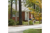 Exterior view of a brick and white siding building partially obscured by trees with green and yellow leaves, a paved driveway, and two white mailboxes in front.