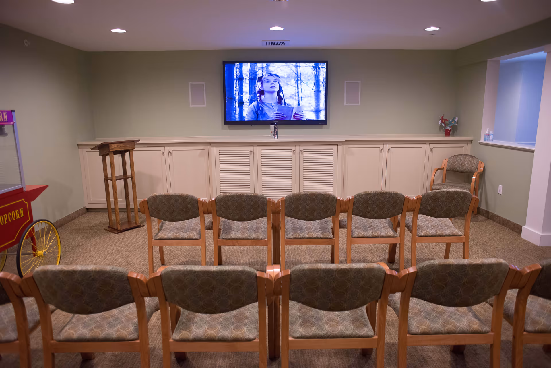 Small screening/activity room with rows of chairs facing a wall-mounted TV and a popcorn cart.