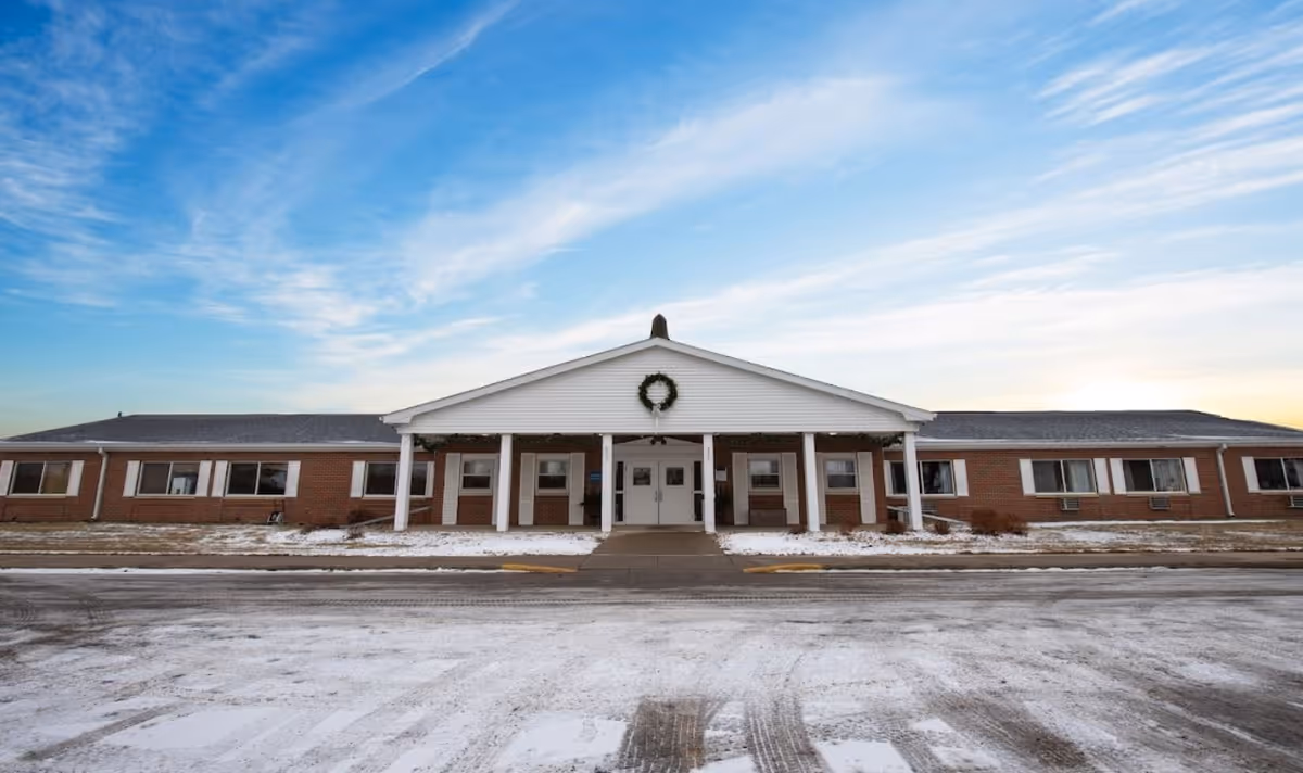 Single-story brick senior living building with a central white portico and wreath, seen across a snowy driveway under a blue sky.