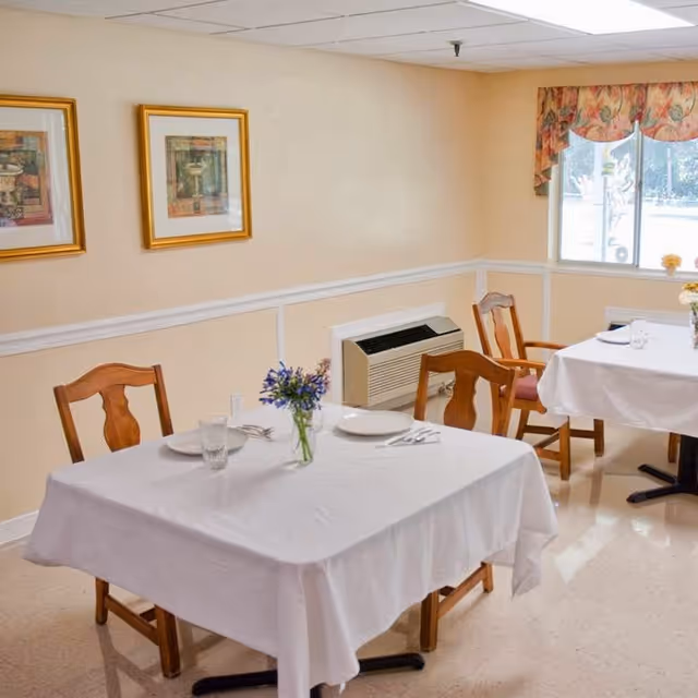 Small dining room with tables draped in white tablecloths set with plates and glasses, wooden chairs, framed artwork on the wall, and a window.