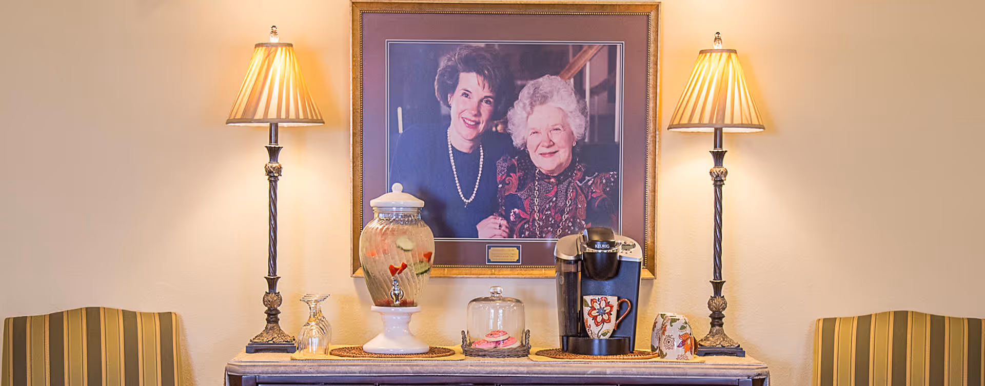 A cozy interior scene with a table holding a beverage dispenser filled with fruit-infused water, a glass jar with cookies, a coffee maker with a floral mug, and two table lamps with beige shades. Above the table is a framed portrait of two smiling elderly women. Two striped armchairs flank the table on either side.