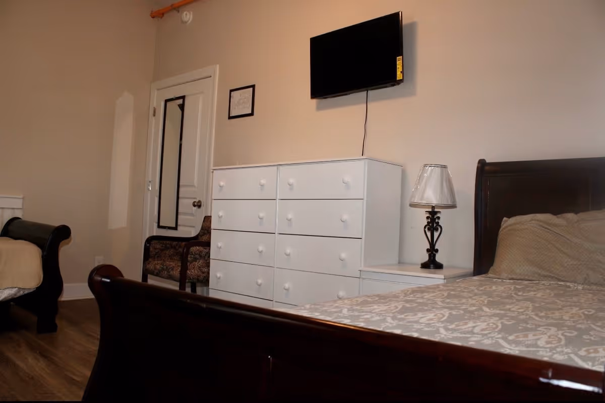 A bedroom with two dark wooden beds, a white dresser with multiple drawers, a small bedside table with a lamp, a wall-mounted flat screen TV, a patterned armchair, and a door with a full-length mirror attached. The room has light-colored walls and wooden flooring.