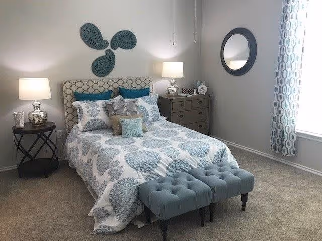 A neatly made bed with a patterned blue and white comforter and multiple pillows in a bedroom. There are two small blue tufted benches at the foot of the bed. On the left side, there is a round side table with a lamp and decorative items. On the right side, there is a dresser with a lamp, clock, and other decor. A round mirror hangs on the wall above the dresser, and patterned curtains frame a window on the right.