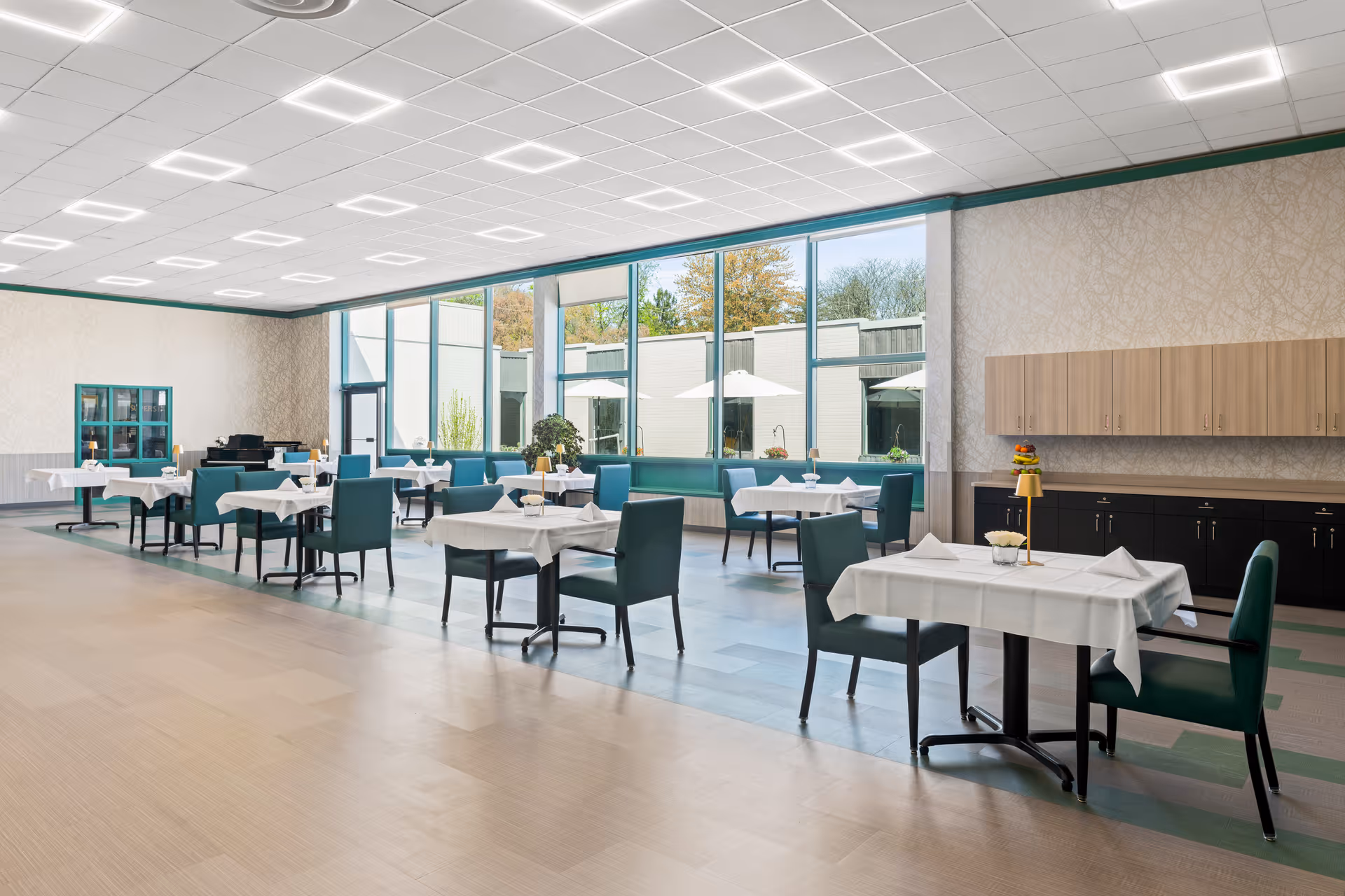 A spacious dining room with multiple tables covered in white tablecloths, each set with napkins and small centerpieces. The room features large windows letting in natural light and offering a view of an outdoor patio area with umbrellas. The walls have light-colored wallpaper, and there are cabinets along one side of the room. The ceiling has modern square light fixtures.