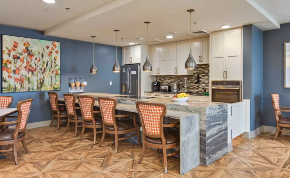 Communal dining area with a stone-topped island, pendant lights, multiple chairs, and a kitchen in the background.