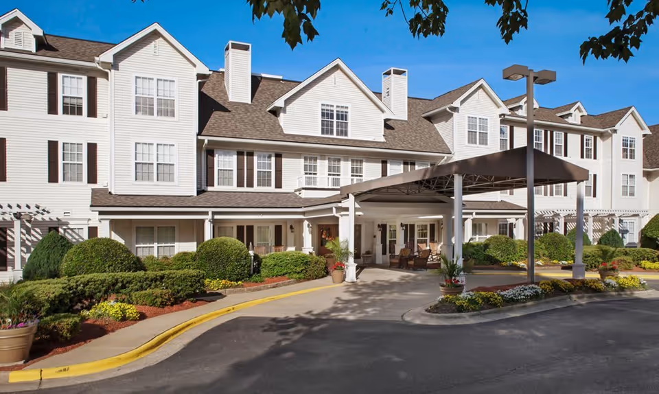 Exterior front view of a multi-story senior living facility building with white siding and brown shutters. The entrance has a covered drop-off area with seating and is surrounded by well-maintained landscaping including bushes, flowers, and trees under a clear blue sky.