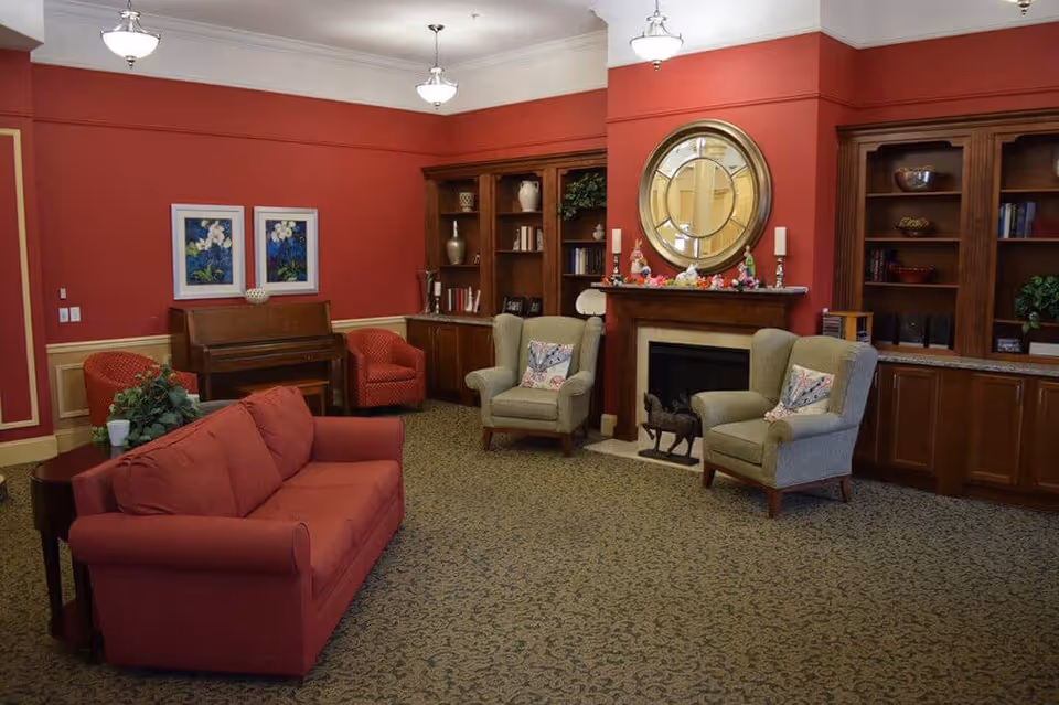 A cozy living room with red walls and carpeted floor featuring a red sofa, two green armchairs with floral pillows, a wooden piano, two red armchairs, built-in wooden bookshelves, a fireplace with a large round mirror above it, and decorative items on the mantel.