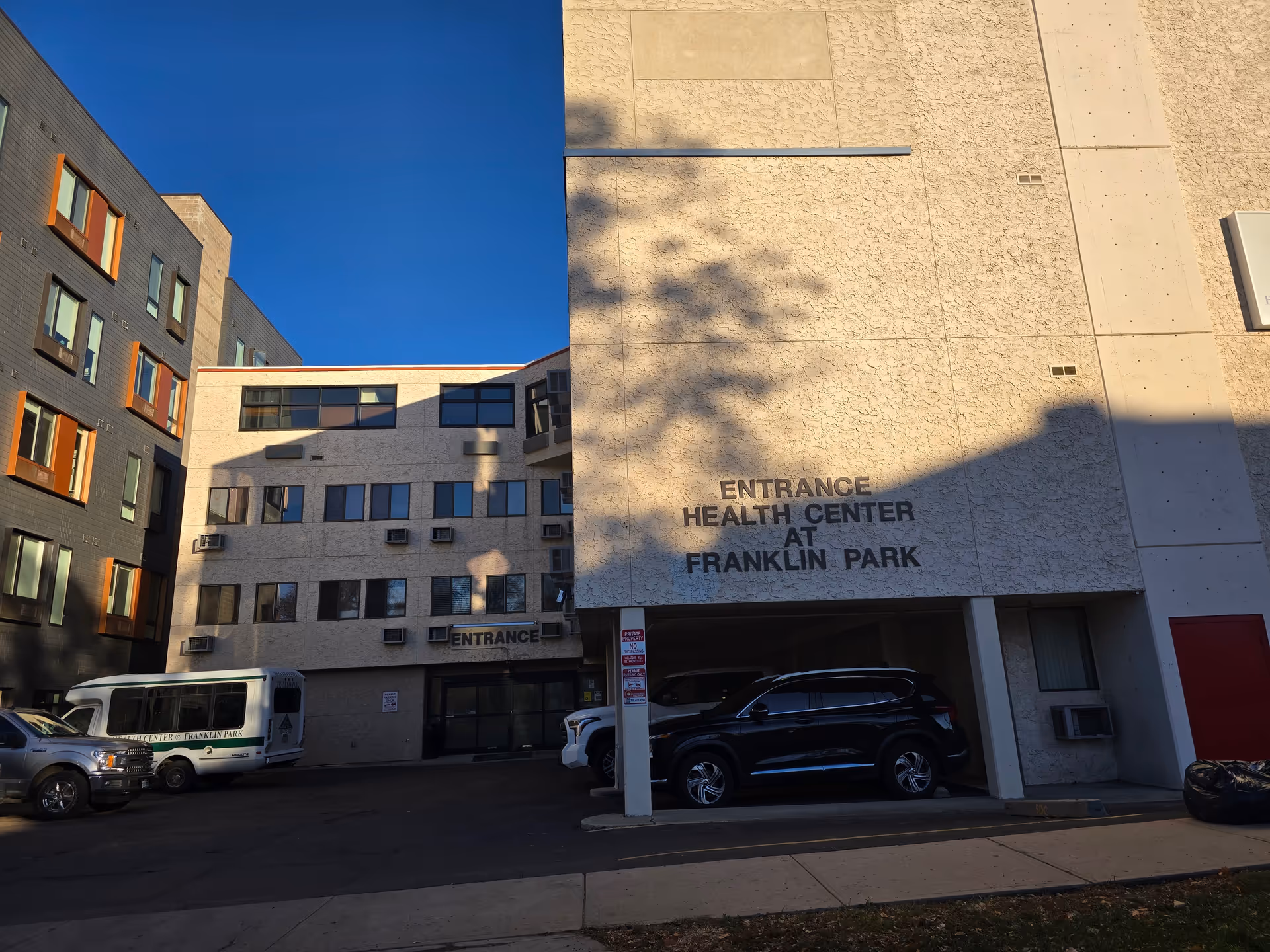 Exterior view of the Health Center at Franklin Park building entrance with several parked vehicles including a van and cars. The building has multiple windows and a textured beige facade with the words 'ENTRANCE HEALTH CENTER AT FRANKLIN PARK' displayed prominently on the wall.